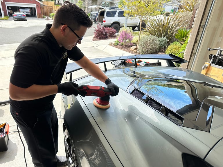 Man in black shirt polishing silver car roof with orbital buffer in driveway