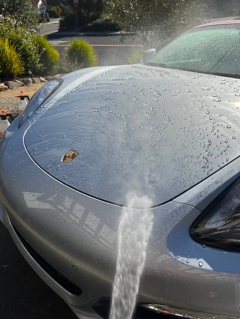 Water streaming onto a silver car during washing, with spray and water droplets covering the hood and windshield